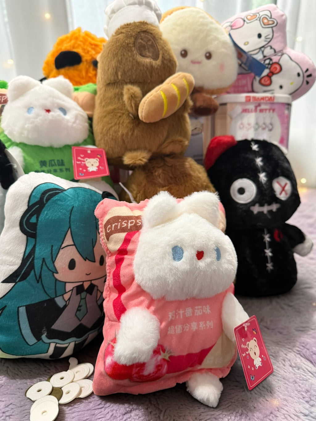 A group of plush toys and character pillows arranged together on a soft purple surface. The front center item is a white plush cat in a pink outfit with a red tag, surrounded by other stuffed toys including a teal anime character pillow, a black stitched figure, a brown bear-like plush, and a Hello Kitty plush in the background.