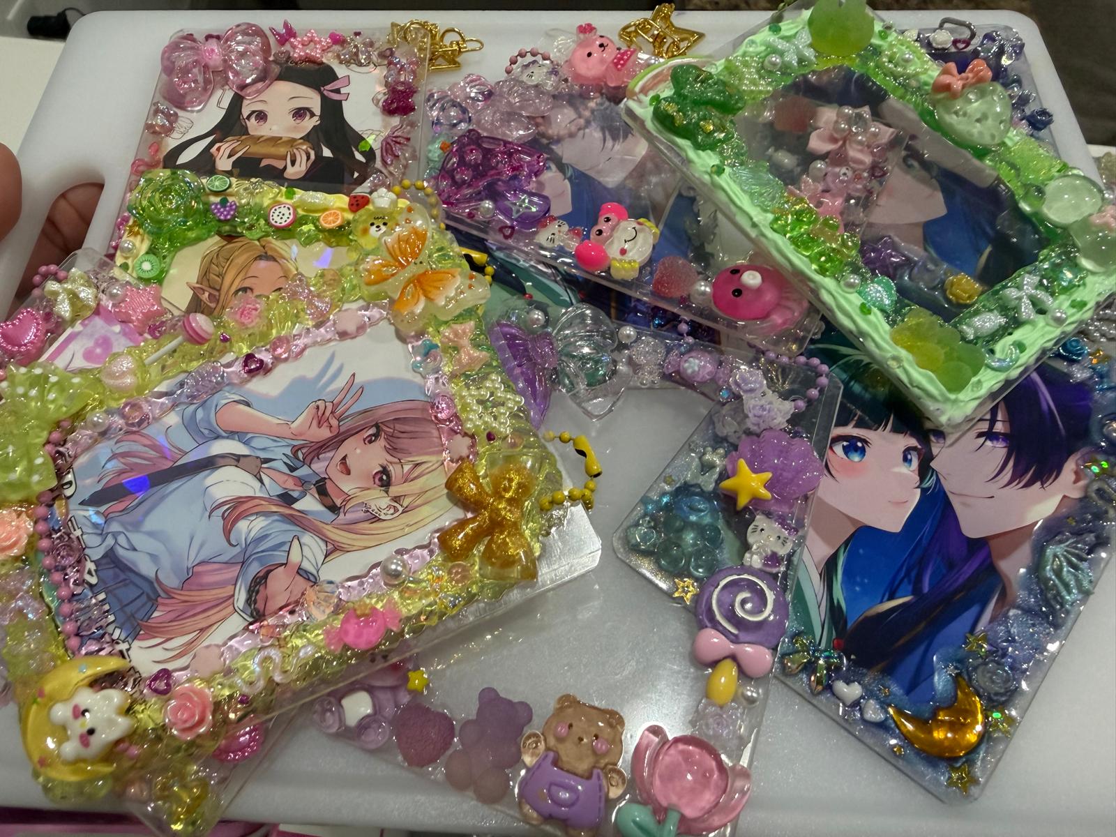 Several decorated transparent phone cases or card holders filled with colorful rhinestones, charms, and anime character prints, arranged on a white surface.
